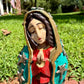 A colorful Milagros Guadalupe  (Virgin Mary) standing on a brick surface outdoors with a grassy yard in the background, close-up of face.