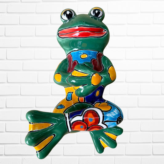 A colorful ceramic frog wall sculpture sitting cross-legged against a white painted brick wall. The frog features a glossy green body with yellow, blue, red, and brown painted patterns, large black-and-white eyes, and a red mouth.