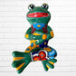 A colorful ceramic frog wall sculpture sitting cross-legged against a white painted brick wall. The frog features a glossy green body with yellow, blue, red, and brown painted patterns, large black-and-white eyes, and a red mouth.