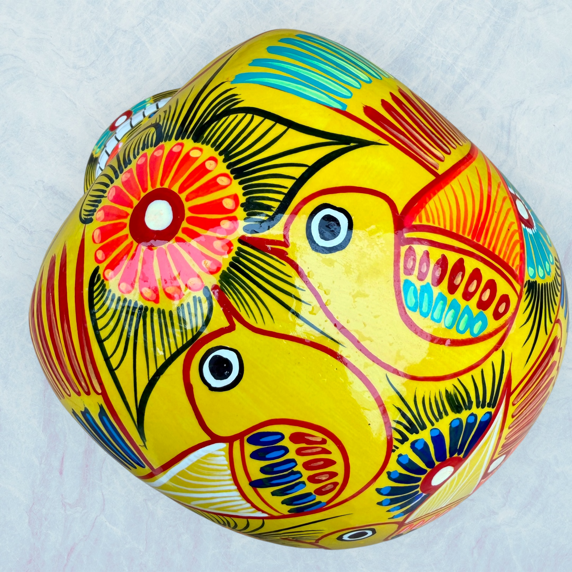 A vibrant yellow sugar skull decorated with colorful floral and geometric patterns in blue, red, black, and white, set against a light blue textured background, top view of skull.