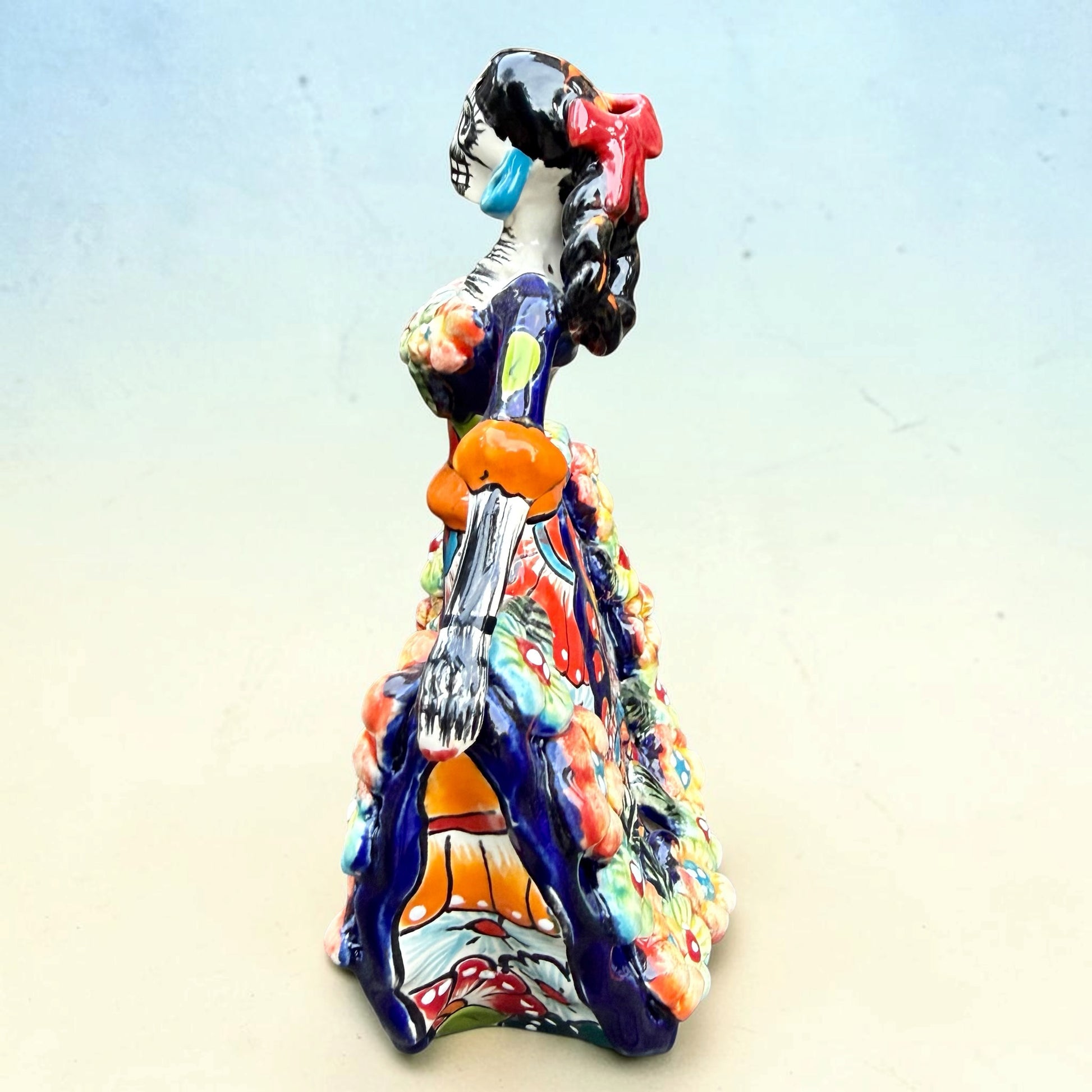 A colorful talavera Day of the Dead (Día de los Muertos) dancing skeleton ballerina figurine. The figurine features black hair in braids wearing a vibrant multicolor dress with floral patterns and orange accents, set against a light blue and beige gradient background, right side view.