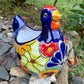 A colorful ceramic chicken-shaped planter with a glossy hand-painted finish, placed outdoors on a bed of small gray rocks and greenery.