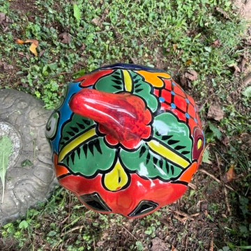 A hand-painted talavera halloween  jack-o'-lantern candle holder in red, green, black, and orange, displayed on a stone pedestal in an outdoor garden setting top of pumpkin view.