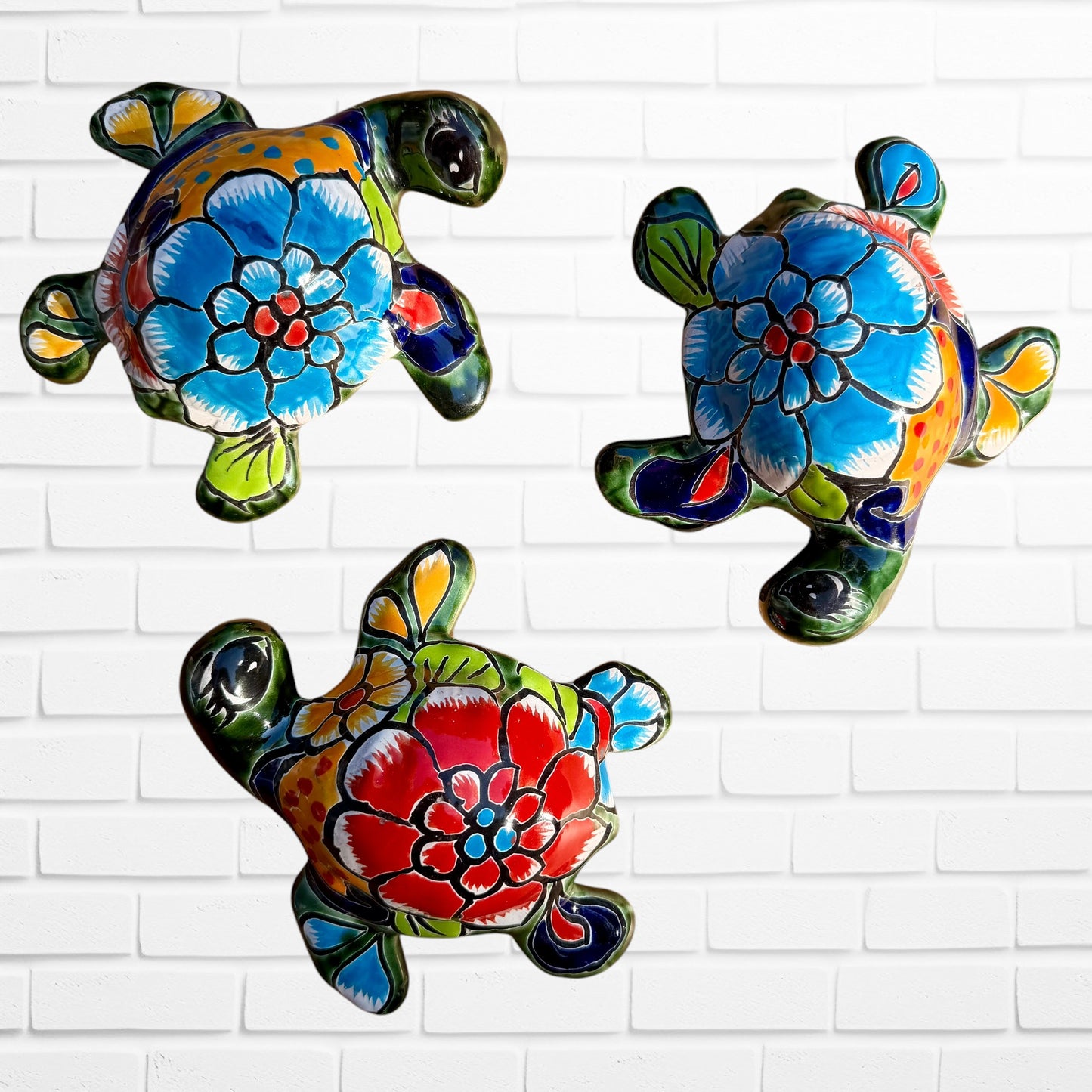 Three colorful hand-painted talavera ceramic sea turtles with floral patterns on their shells, displayed against a white brick wall background.
