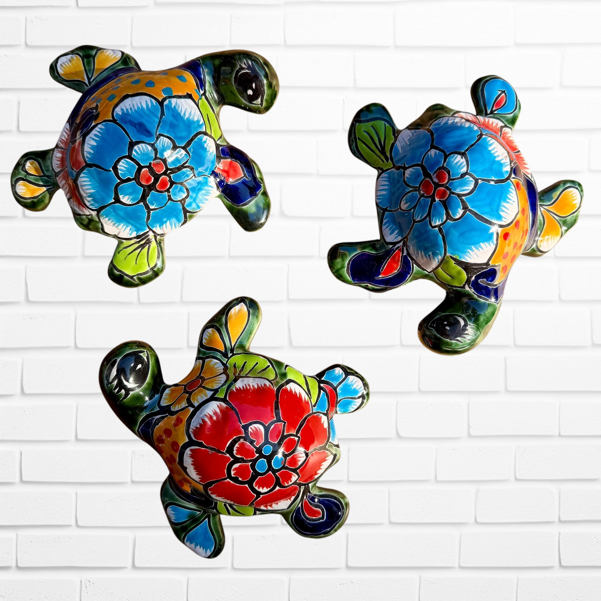 Three colorful hand-painted talavera ceramic sea turtles with floral patterns on their shells, displayed against a white brick wall background.