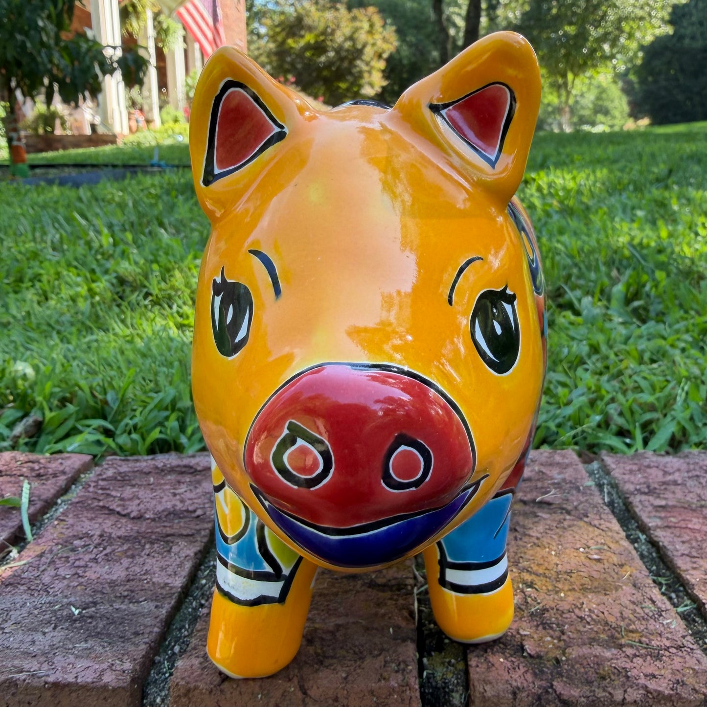 A colorful hand-painted yellow Talavera pig figurine standing on a brick surface with a blurred outdoor garden background, front face view.