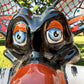 A colorful Talavera dog statue holding a big white bone, photographed outdoors on grass with trees in the background. The dog has a glossy black body, blue eyes, an orange snout, and multicolored patterned ears. Close up of the puppies big blue eyes.