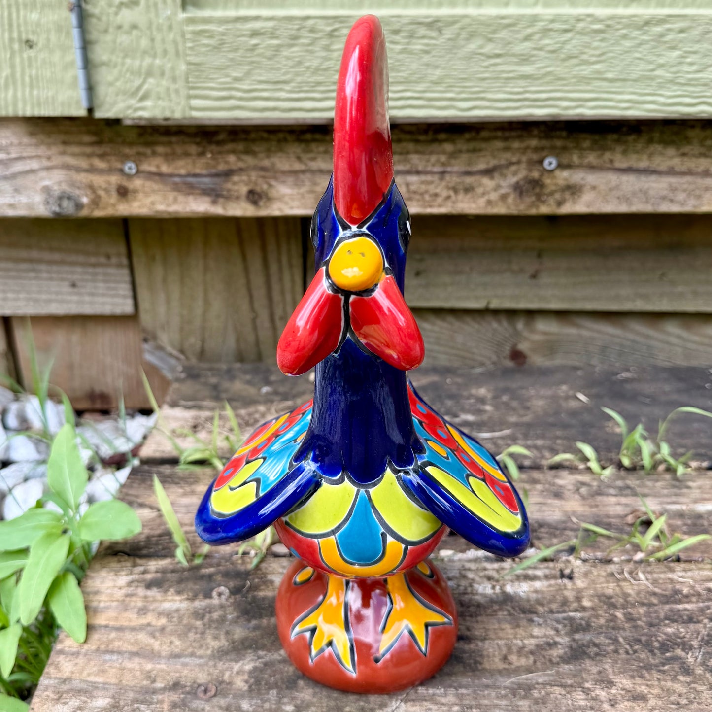 A colorful ceramic rooster figurine standing on a weathered wooden surface outdoors, with a green wooden wall behind it. The rooster is decorated with bright red, yellow, blue, and orange painted details, photo of front bore.