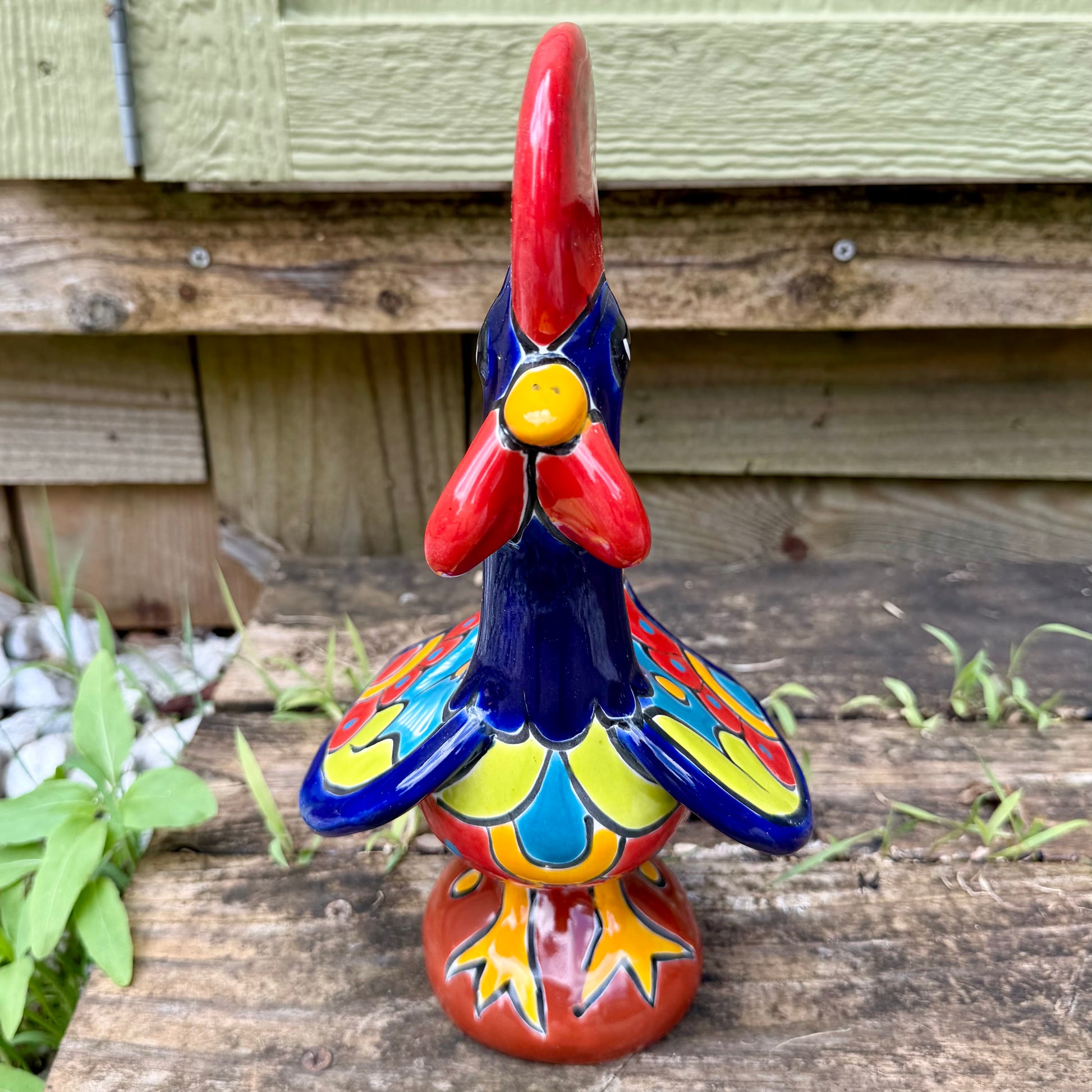 A colorful ceramic rooster figurine standing on a weathered wooden surface outdoors, with a green wooden wall behind it. The rooster is decorated with bright red, yellow, blue, and orange painted details, photo of front bore.