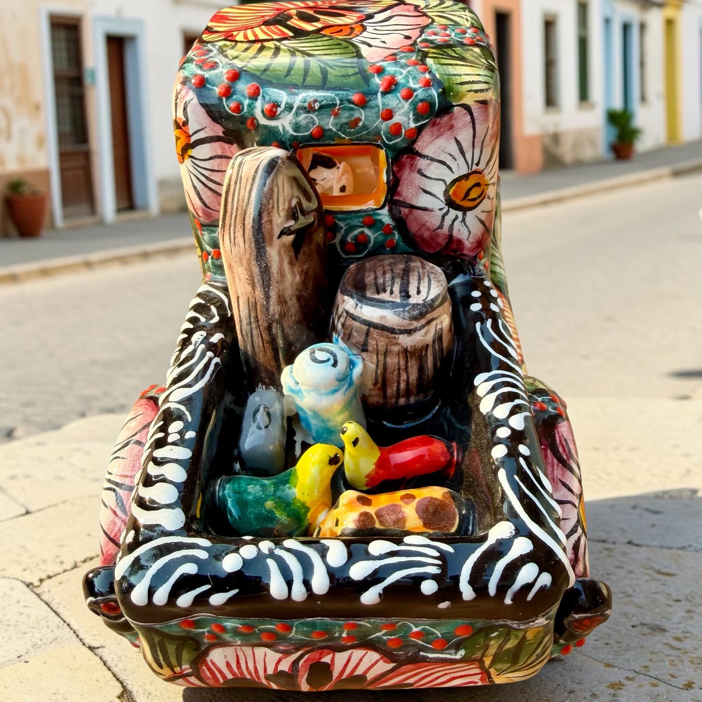 A decorative Talavera day of the dead pick up truck figurine two skeleton passengers. The truck is vibrant, with a hand-painted tropical floral pattern, photographed outdoors on a stone-paved street. This is the back view of the pick up truck, Showing farm animals and a coffin.