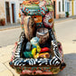 A decorative Talavera day of the dead pick up truck figurine two skeleton passengers. The truck is vibrant, with a hand-painted tropical floral pattern, photographed outdoors on a stone-paved street. This is the back view of the pick up truck, Showing farm animals and a coffin.