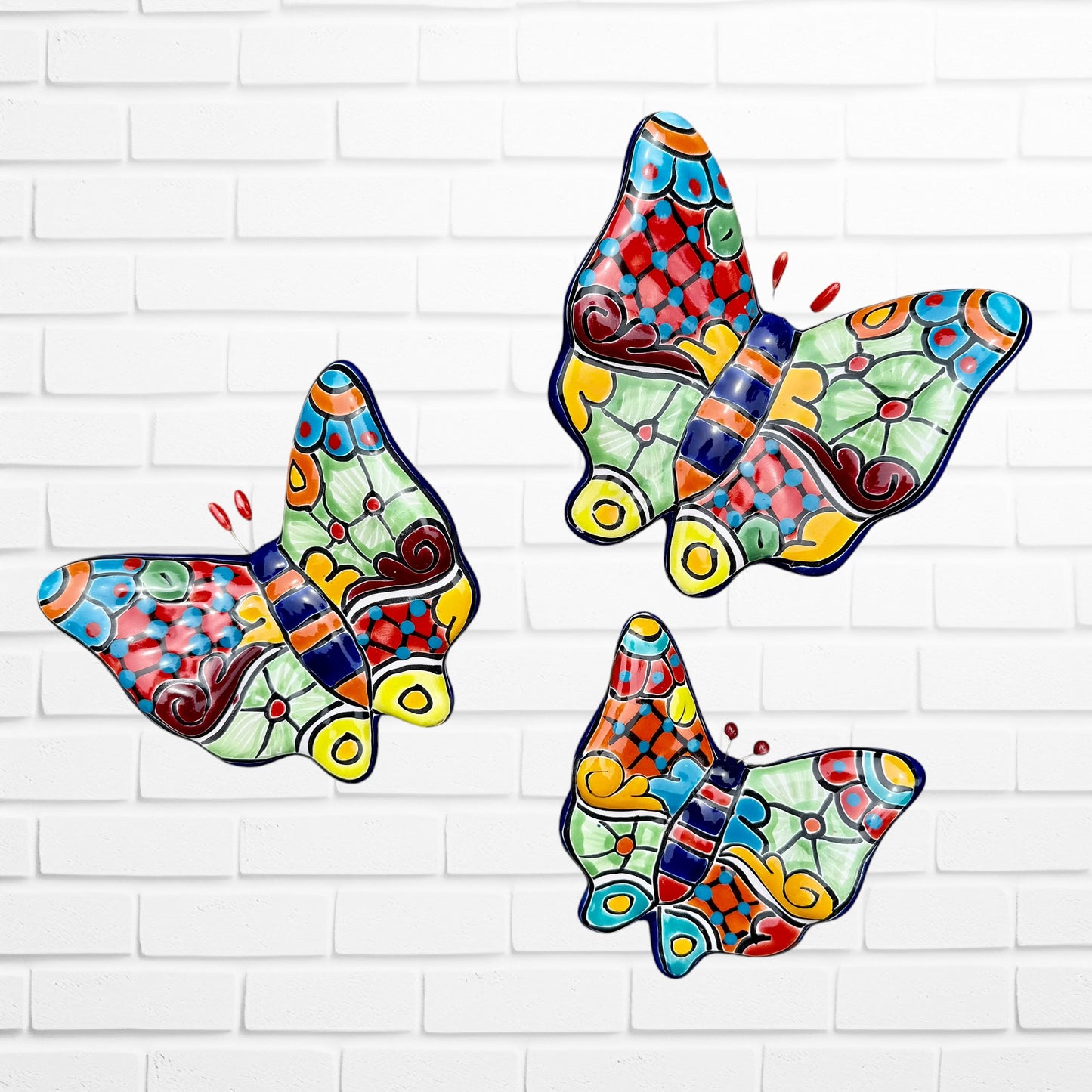 Three colorful ceramic Talavera butterfly wall art pieces with orange, blue, green, yellow, and purple mosaic patterns, displayed against a white brick wall.