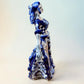 A Day of the Dead style talavera figurine of a skeleton woman in a wide floral skirt, painted in deep blue, black, white, and light blue on a soft beige-to-blue gradient background. Left side view of ballerina.