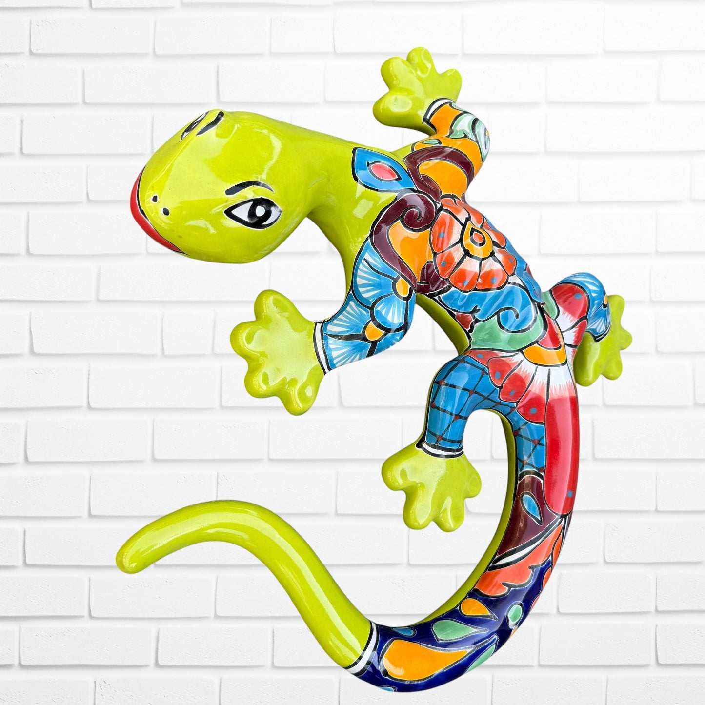 A colorful decorative lizard Talavera wall sculpture with a hand-painted floral and geometric pattern, mounted against a white brick wall, left side view.
