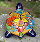 A glossy ceramic turtle figurine with a vibrant hand-painted patterned shell and a deep blue head and legs, resting on a gray stone surface with a blurred green garden background, back view of Shell.