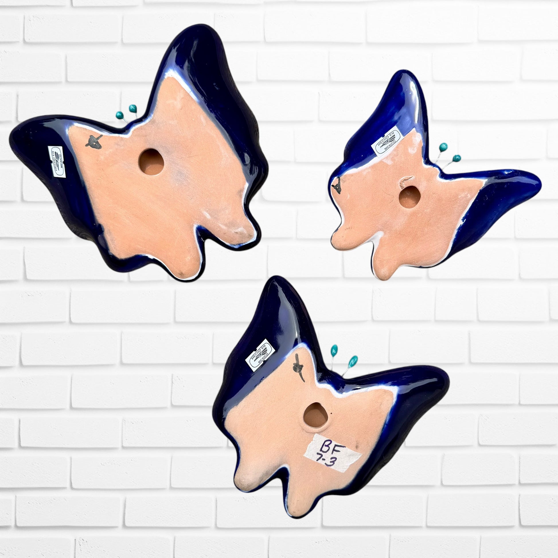 Three colorful ceramic Talavera butterfly wall art pieces with orange, blue, green, yellow, and purple mosaic patterns, displayed against a white brick wall. Back view with hanging holes.
