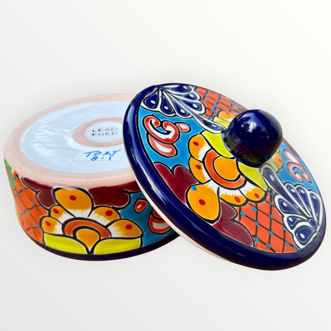 A hand-painted Talavera ceramic lidded container with a colorful Mexican-style floral pattern in blue, orange, yellow, red, and white, featuring a glossy dark blue lid knob, exterior side view. 