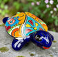 A glossy ceramic turtle figurine with a vibrant hand-painted patterned shell and a deep blue head and legs, resting on a gray stone surface with a blurred green garden background.