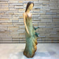 Loretta Bertoli faceless female sculpture wearing a long sleeveless gown, standing against a stone tile wall. The dress has a distressed, textured finish in muted teal, brown, and beige tones, with  hummingbird figures attached near the lower skirt.