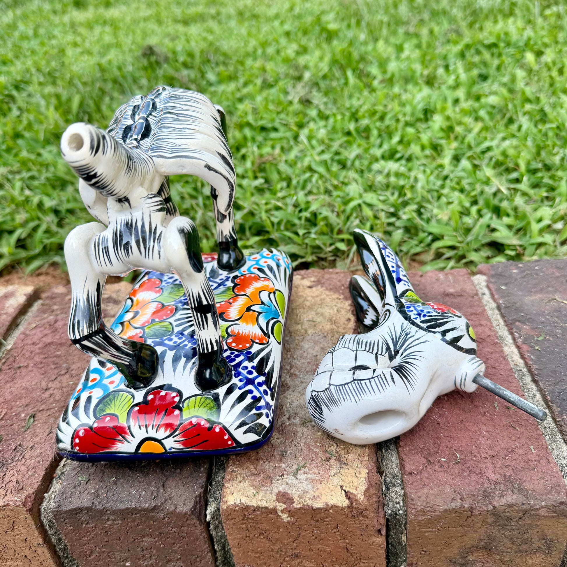 A colorful ceramic donkey Catrina figurine with a skeletal white-and-black design and a floral base, photographed outdoors on a brick surface with a blurred green lawn and house in the background. Photo Showing donkey with his head off.