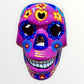 A hand-painted ceramic sugar skull mask in vibrant magenta pink, decorated with colorful floral and geometric patterns in blue, yellow, red, and white, set against a plain white background Front face view.