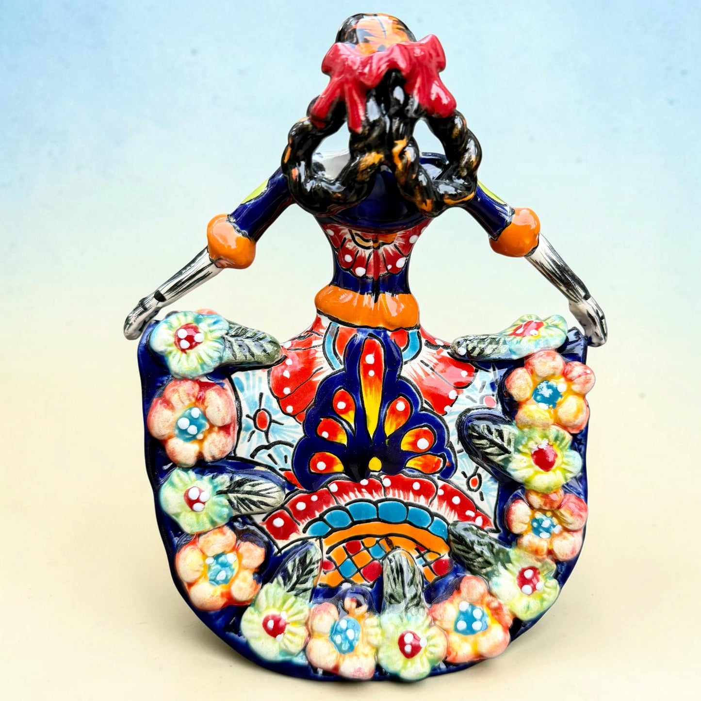 A colorful talavera Day of the Dead (Día de los Muertos) dancing skeleton ballerina figurine. The figurine features black hair in braids wearing a vibrant multicolor dress with floral patterns and orange accents, set against a light blue and beige gradient background, back view.