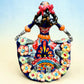 A colorful talavera Day of the Dead (Día de los Muertos) dancing skeleton ballerina figurine. The figurine features black hair in braids wearing a vibrant multicolor dress with floral patterns and orange accents, set against a light blue and beige gradient background, back view.