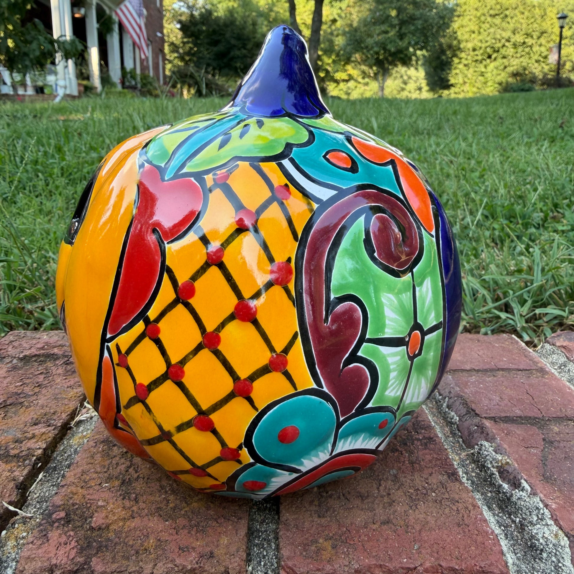A Talavera jack-o'-lantern pumpkin decoration with a carved face, sitting on a brick surface in an outdoor yard with grass and trees in the background. Right side view  of pumpkin.