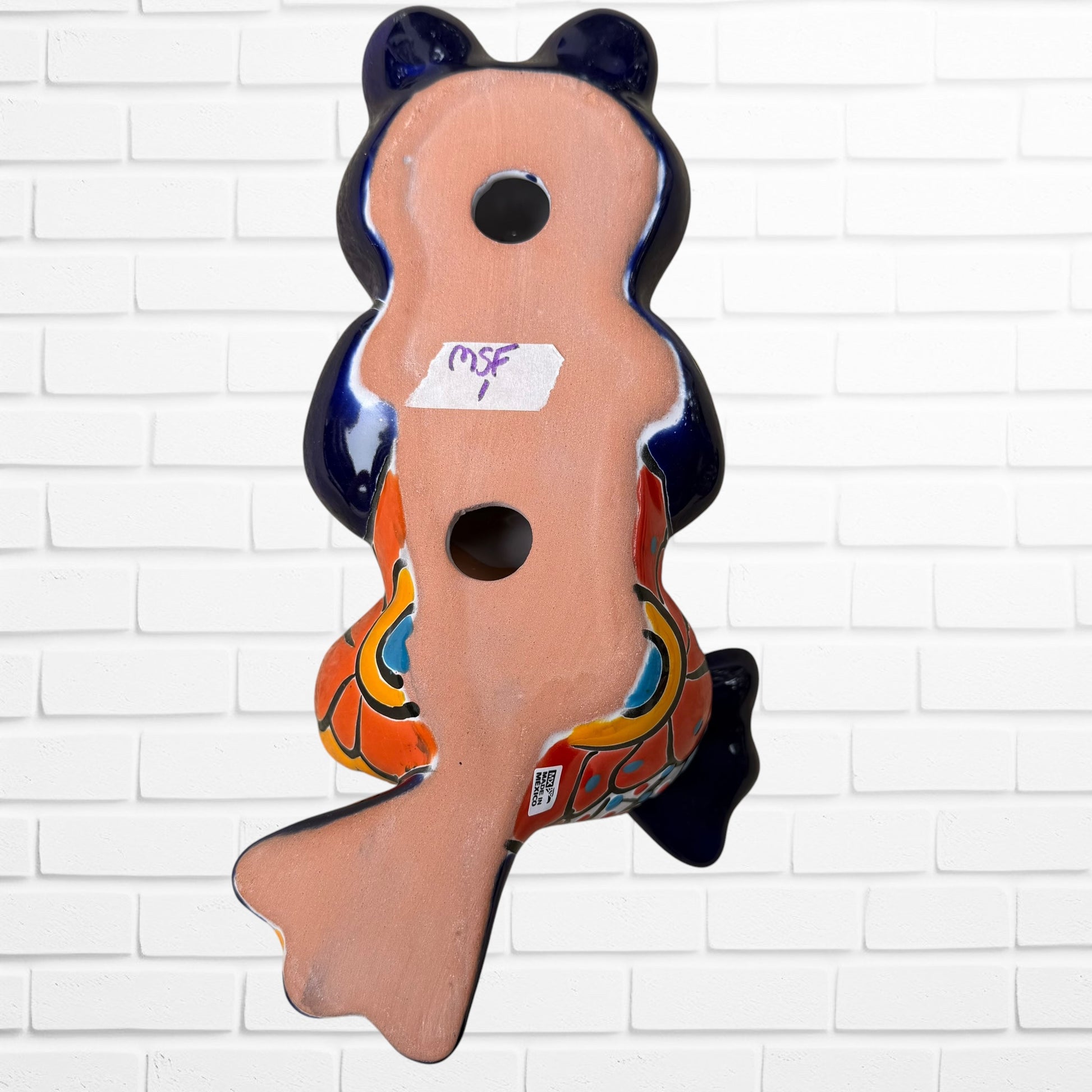 A colorful ceramic frog figurine sitting with arms crossed, decorated with vibrant hand-painted patterns in blue, yellow, red, orange, and light blue, set back view with hanging holes. 