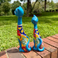 Two colorful ceramic cat figurines sitting on a brick path in a grassy park setting. This is their right side view.