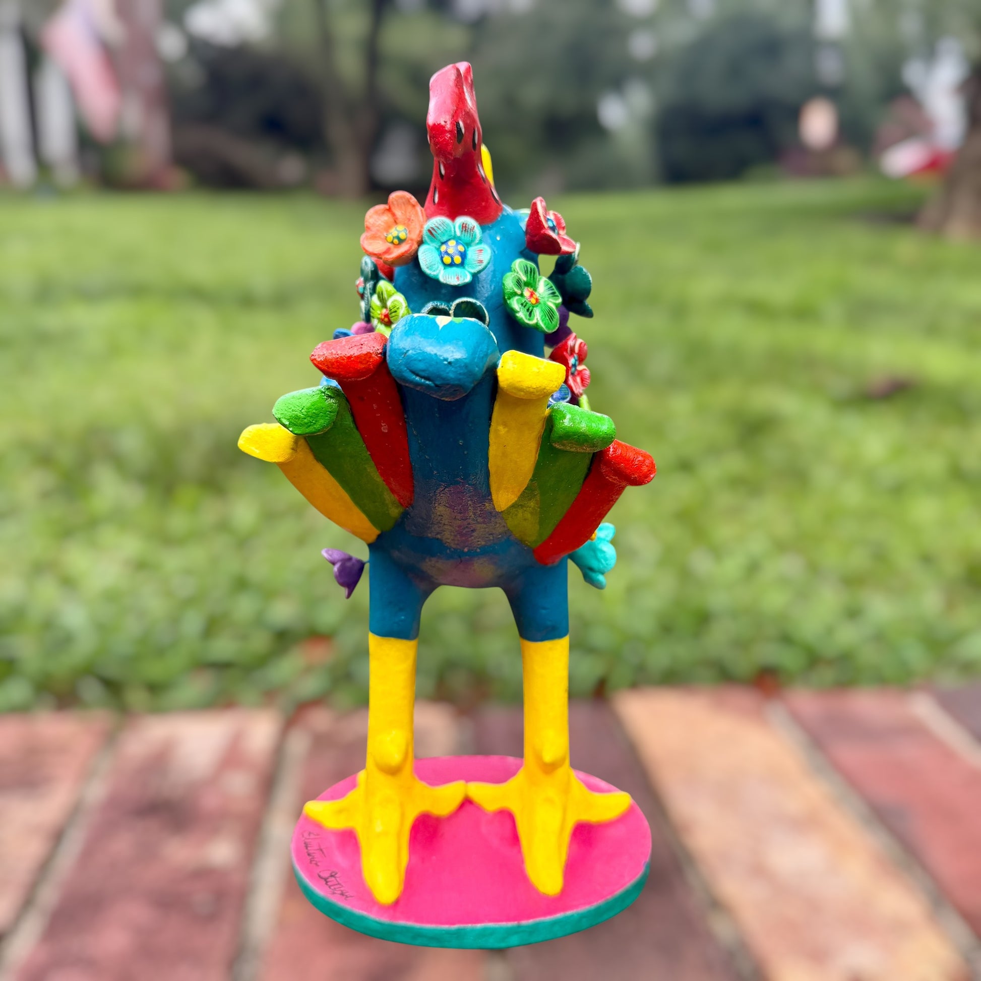 A colorful decorative rooster folk art sculpture with a blue body, yellow legs and beak, and many bright flower ornaments, standing on a pink circular base outdoors on a brick path with a blurred green lawn behind it. Back tail view of rooster.