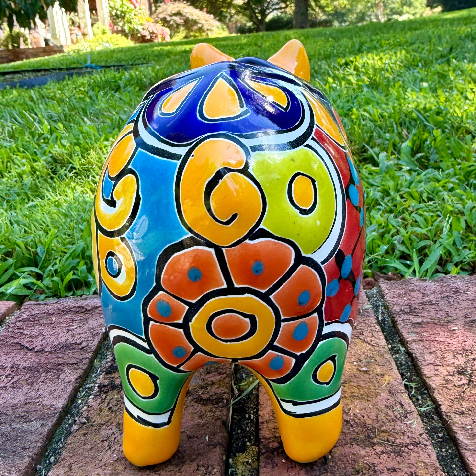 A colorful hand-painted yellow Talavera pig figurine standing on a brick surface with a blurred outdoor garden background, back tail view.