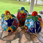 Three colorful hand-painted Talavera jack-o'-lantern pumpkins with multi-color displayed on a stone patio with a blurred green lawn background, back side view.