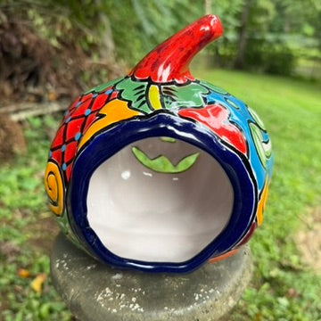 A hand-painted talavera halloween  jack-o'-lantern candle holder in red, green, black, and orange, displayed on a stone pedestal in an outdoor garden setting, back opening view.
