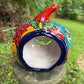 A hand-painted talavera halloween  jack-o'-lantern candle holder in red, green, black, and orange, displayed on a stone pedestal in an outdoor garden setting, back opening view.