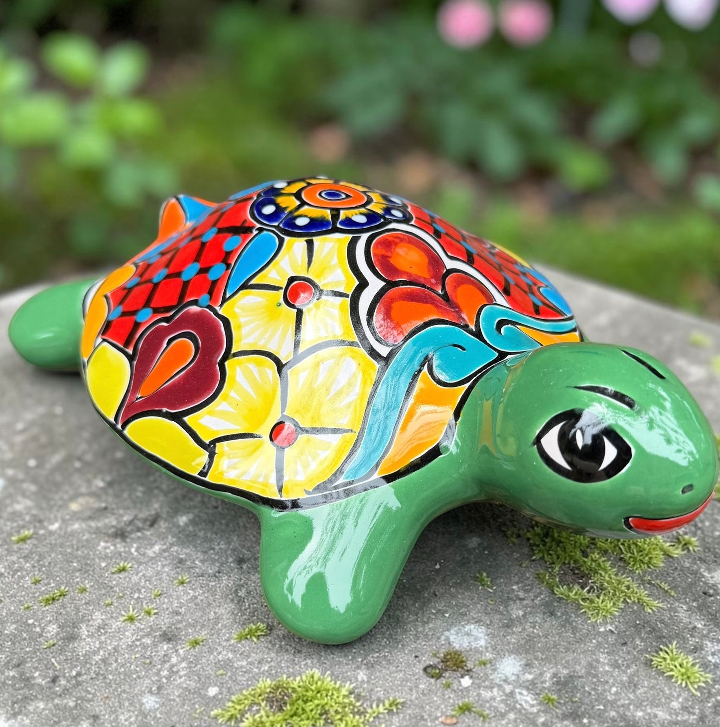 A hand-painted ceramic turtle figurine resting on a gray stone surface with a blurred green garden background. The turtle features a bright green head and legs, and a highly detailed, multicolored shell with yellow, red, orange, blue, and black patterns.