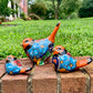 Three hand-painted ceramic bird garden statues in orange, blue, yellow, and black, displayed on a brick ledge in a backyard with a green lawn and trees in the background right side view.