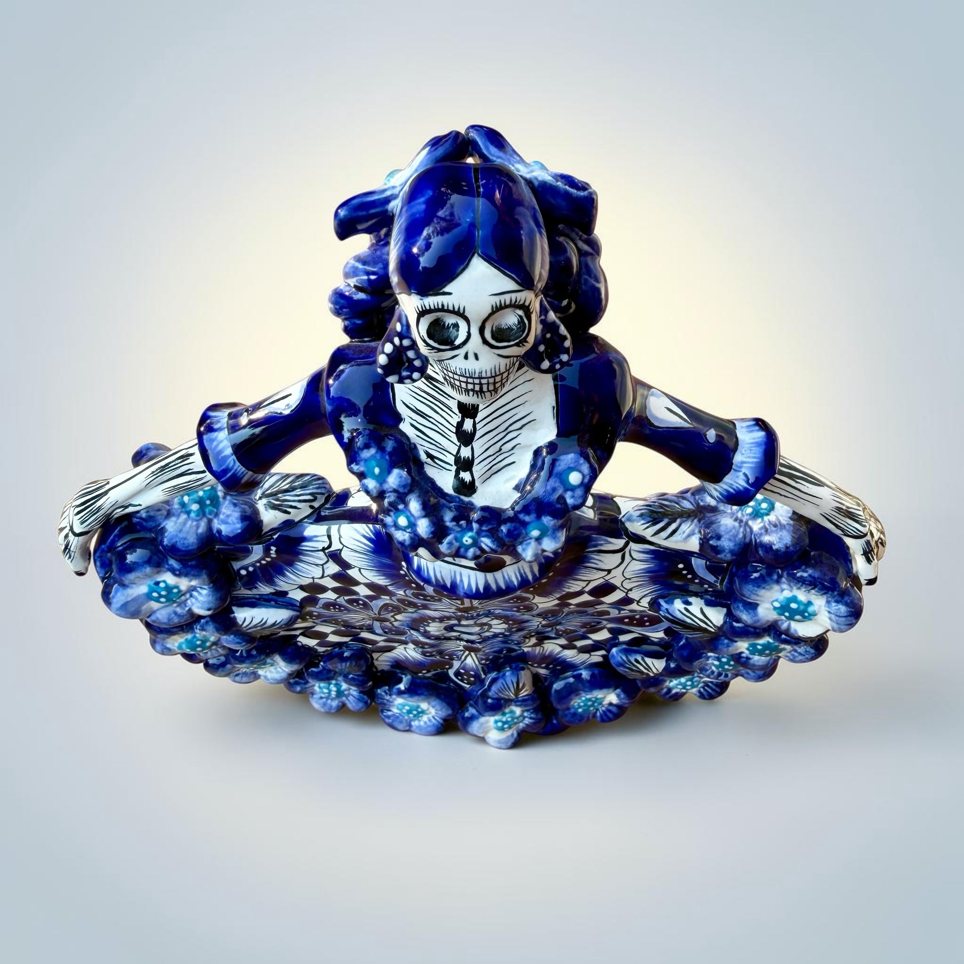 A Day of the Dead style talavera figurine of a skeleton woman in a wide floral skirt, painted in deep blue, black, white, and light blue on a soft beige-to-blue gradient background. Top view of ballerina figurine.