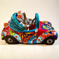 A colorful hand-painted ceramic Mexican-style convertible car figurine with Day of the Dead skeleton characters inside, shown on a neutral beige background. Left side view of the car.
