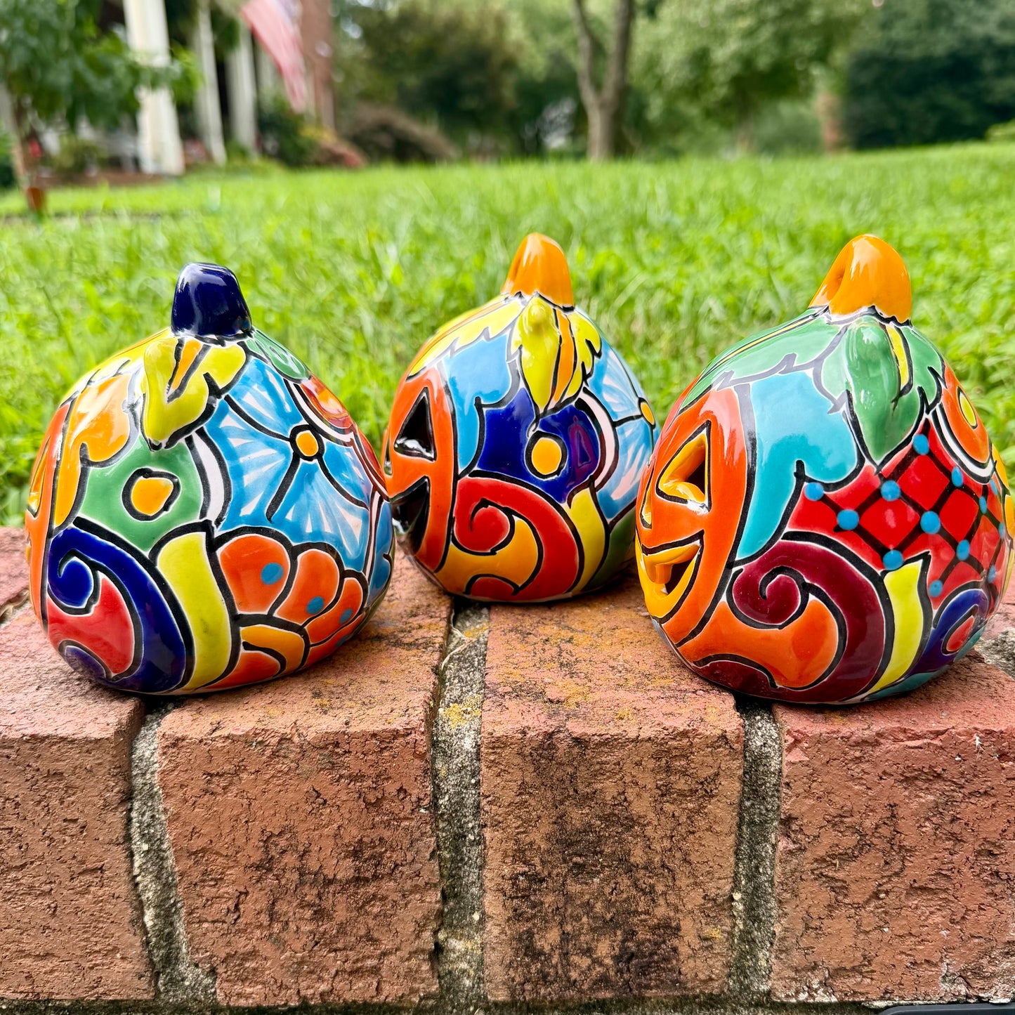 Three hand-painted Talavera ceramic jack-o'-lantern pumpkins with orange faces and yellow, blue, and red green designs, sitting on a brick path in a grassy backyard, left side view.