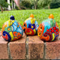Three hand-painted Talavera ceramic jack-o'-lantern pumpkins with orange faces and yellow, blue, and red green designs, sitting on a brick path in a grassy backyard, left side view.