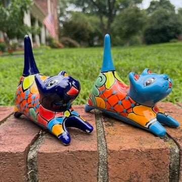 Two colorful hand-painted ceramic cats sitting on a brick ledge with a green lawn and trees in the background. One is cobalt  blue and the other is turquoise, both have traditional multi-color  talavera designs.