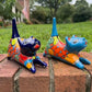 Two colorful hand-painted ceramic cats sitting on a brick ledge with a green lawn and trees in the background. One is cobalt  blue and the other is turquoise, both have traditional multi-color  talavera designs.