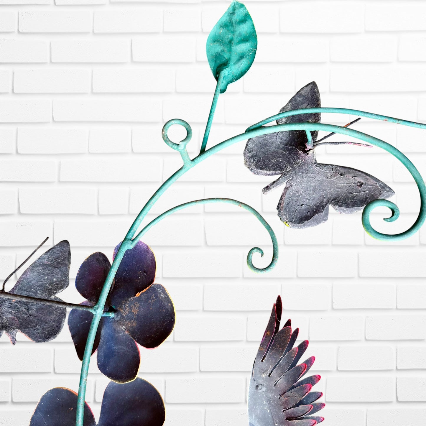 Decorative metal wall art with butterfly, hummingbird  and  flowers with  leaf design against a white brick wall back view hanging loop
