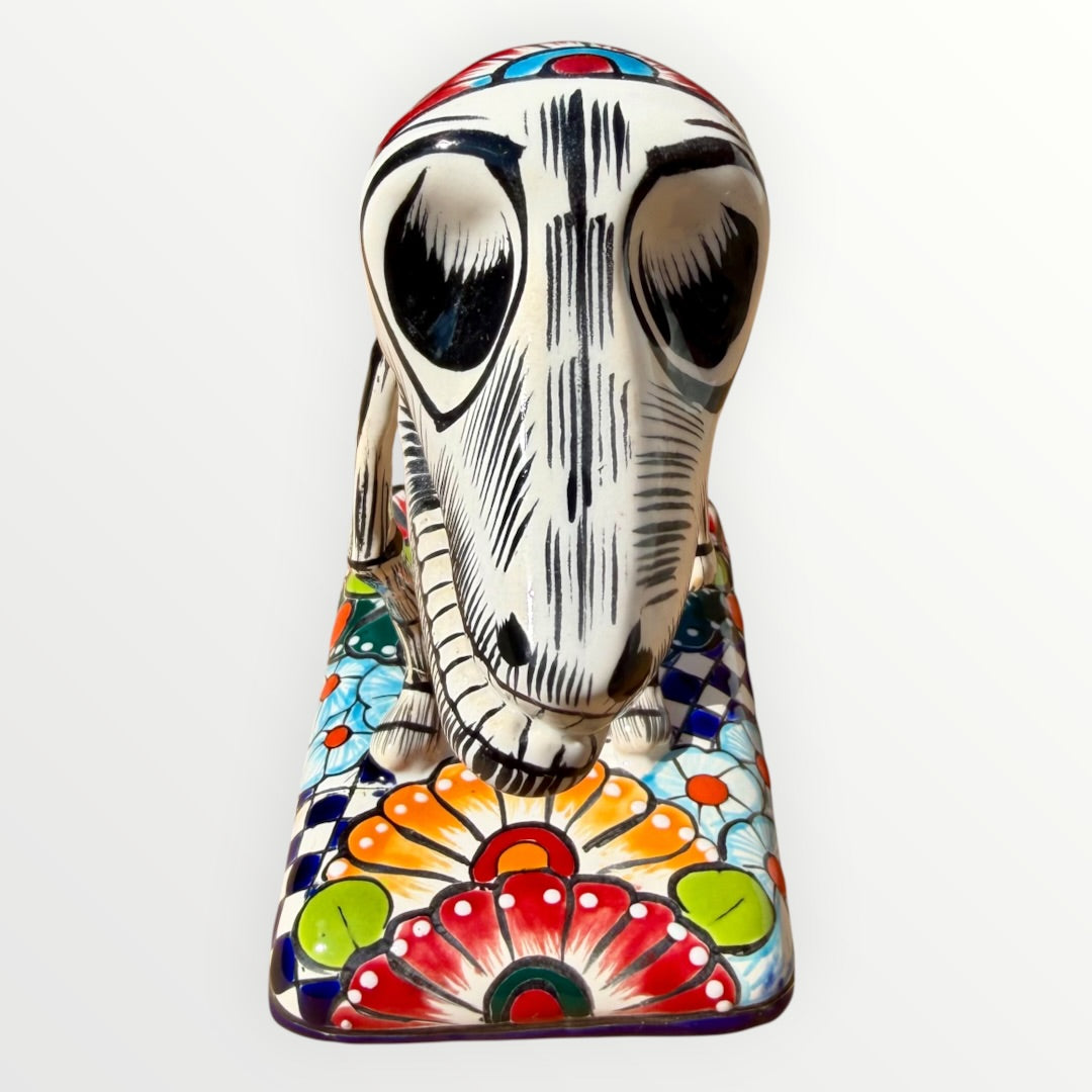 A ceramic Mexican-style skeleton dog figurine standing on a colorful floral base against a plain white background. Front face close up of catrina dog.