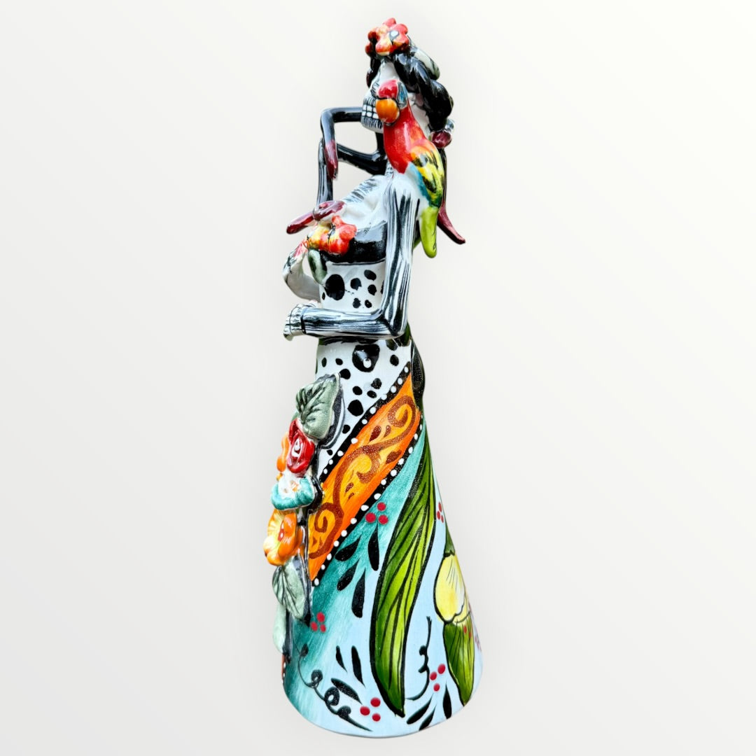 A Day of the Dead Catrina of a Frida skeleton doll holding a black cat, with a red parrot on her shoulder and colorful flowers around the top of her dress. Right side of doll showing a red parrot.