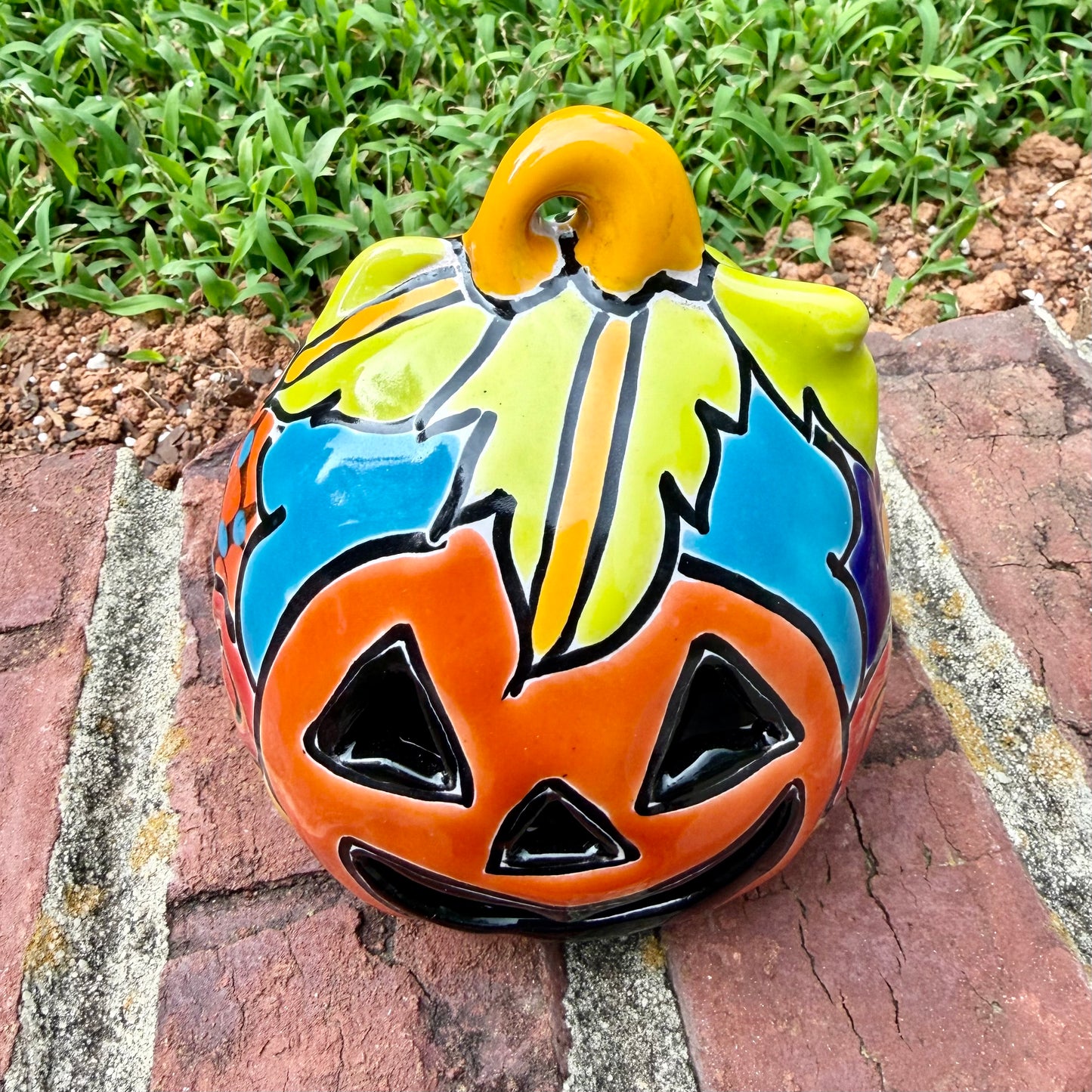 One hand-painted Talavera ceramic jack-o'-lantern pumpkins with orange faces and yellow, blue, and red green designs, sitting on a brick path in a grassy backyard, top stem view.