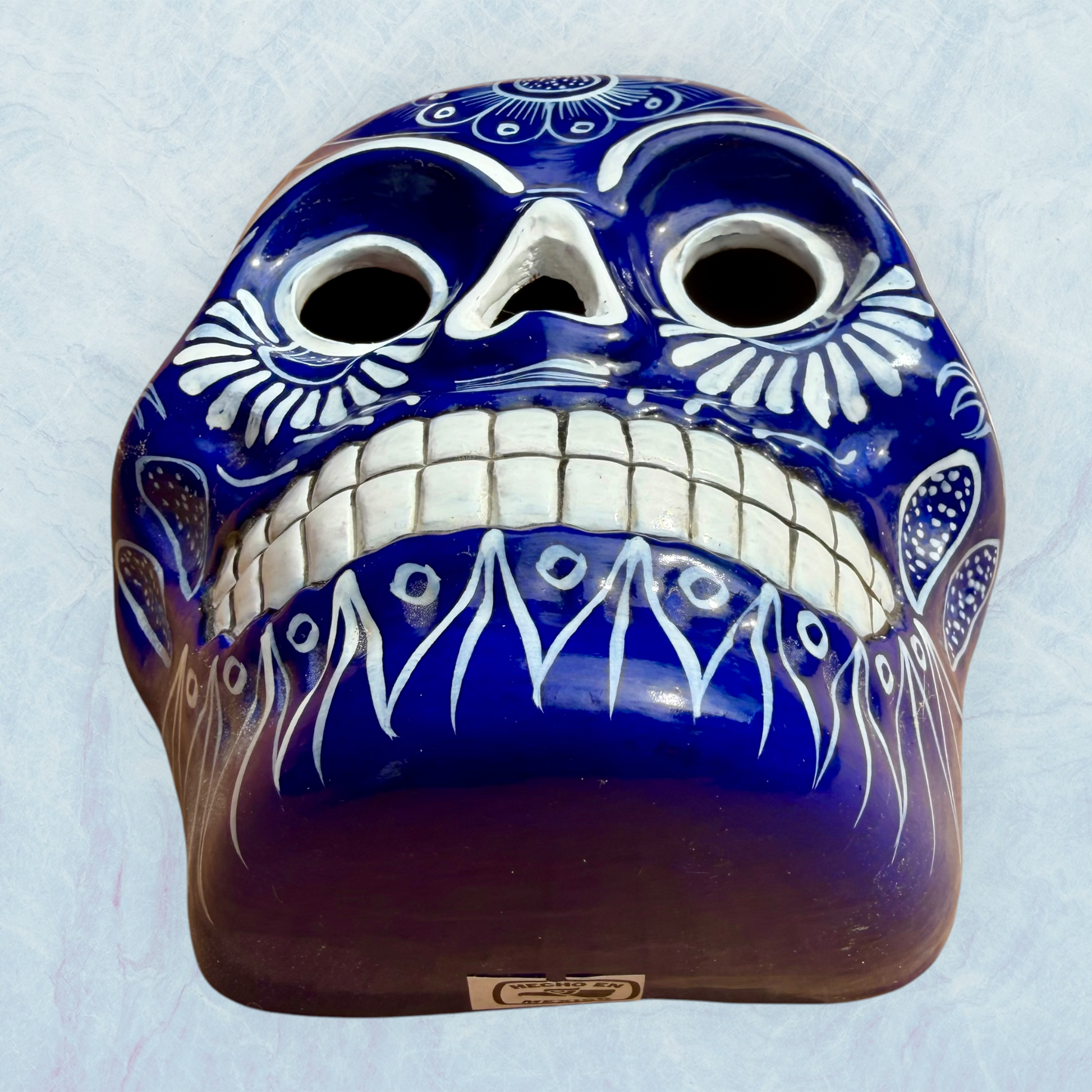 A clay, blue with white designs, Mexican-style skeleton skull face mask hanging on a soft blue surface, bottom chin view.