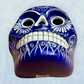 A clay, blue with white designs, Mexican-style skeleton skull face mask hanging on a soft blue surface, bottom chin view.