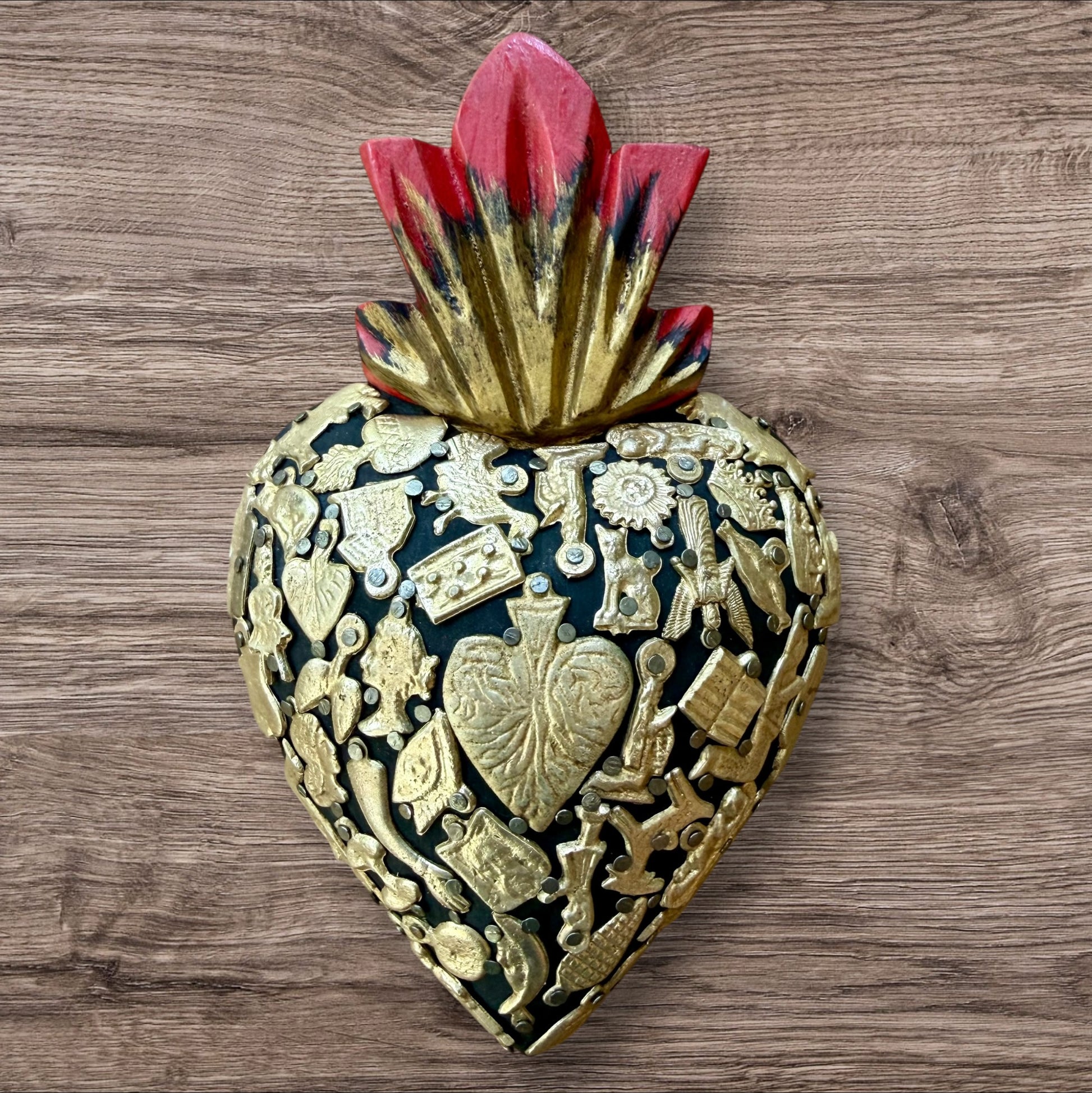 A Mexican Milagros medium heart in black with a gold and red flaming top, covered in antiqued golden charms. 